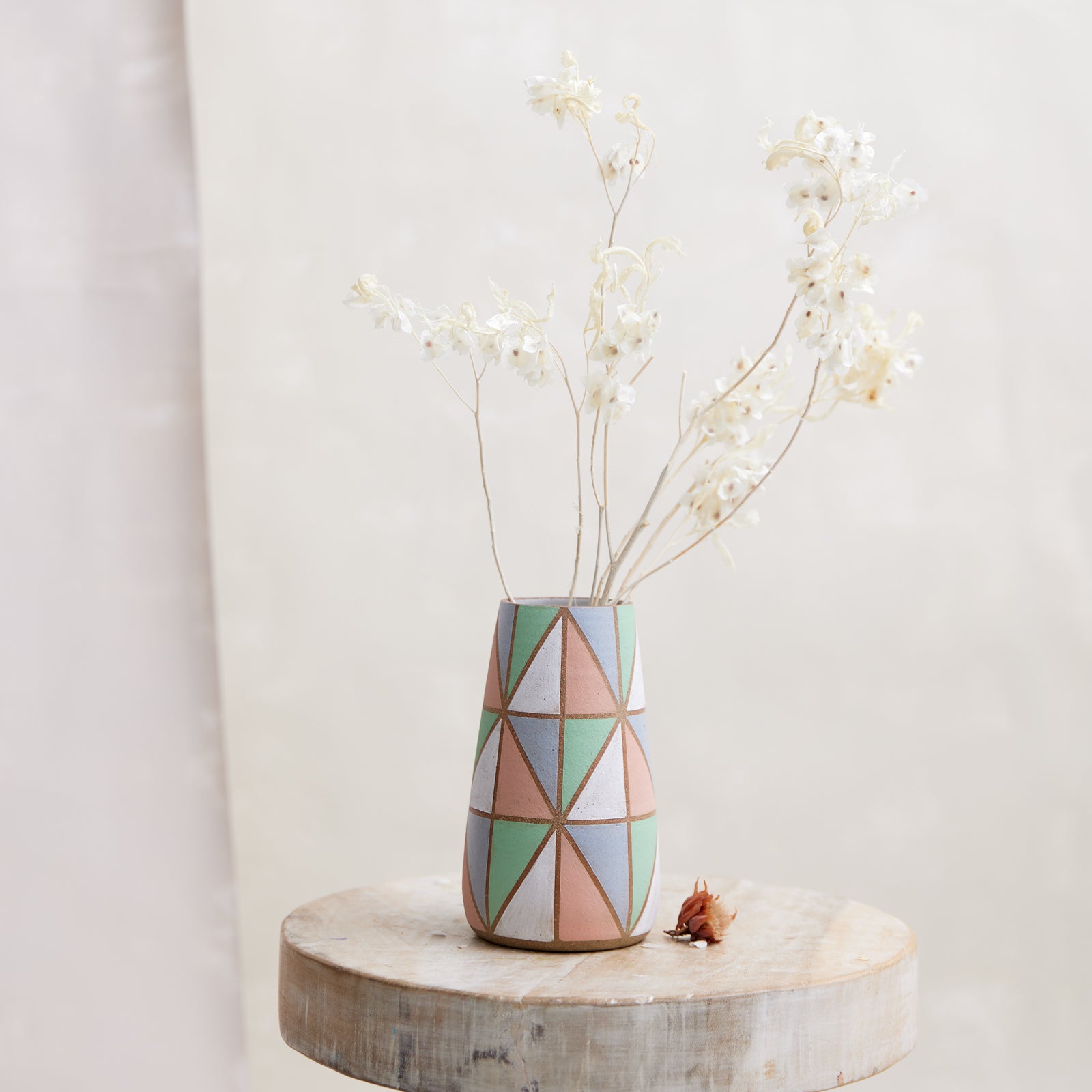 A front view of the Geometric Handmade Ceramic Teardrop Vase in pastel-coloured glazes. This handmade vase holds coastal dried flowers and sits on a wooden stool.