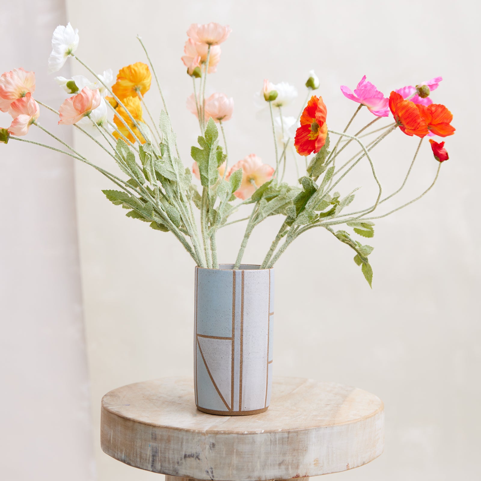 A front view of the Geometric Cylindrical Handmade Ceramic Vase in a blue, white and grey glaze. The handmade vase sits on a wooden stool in a coastal-styled setting. The ceramic vase displays fresh flowers.