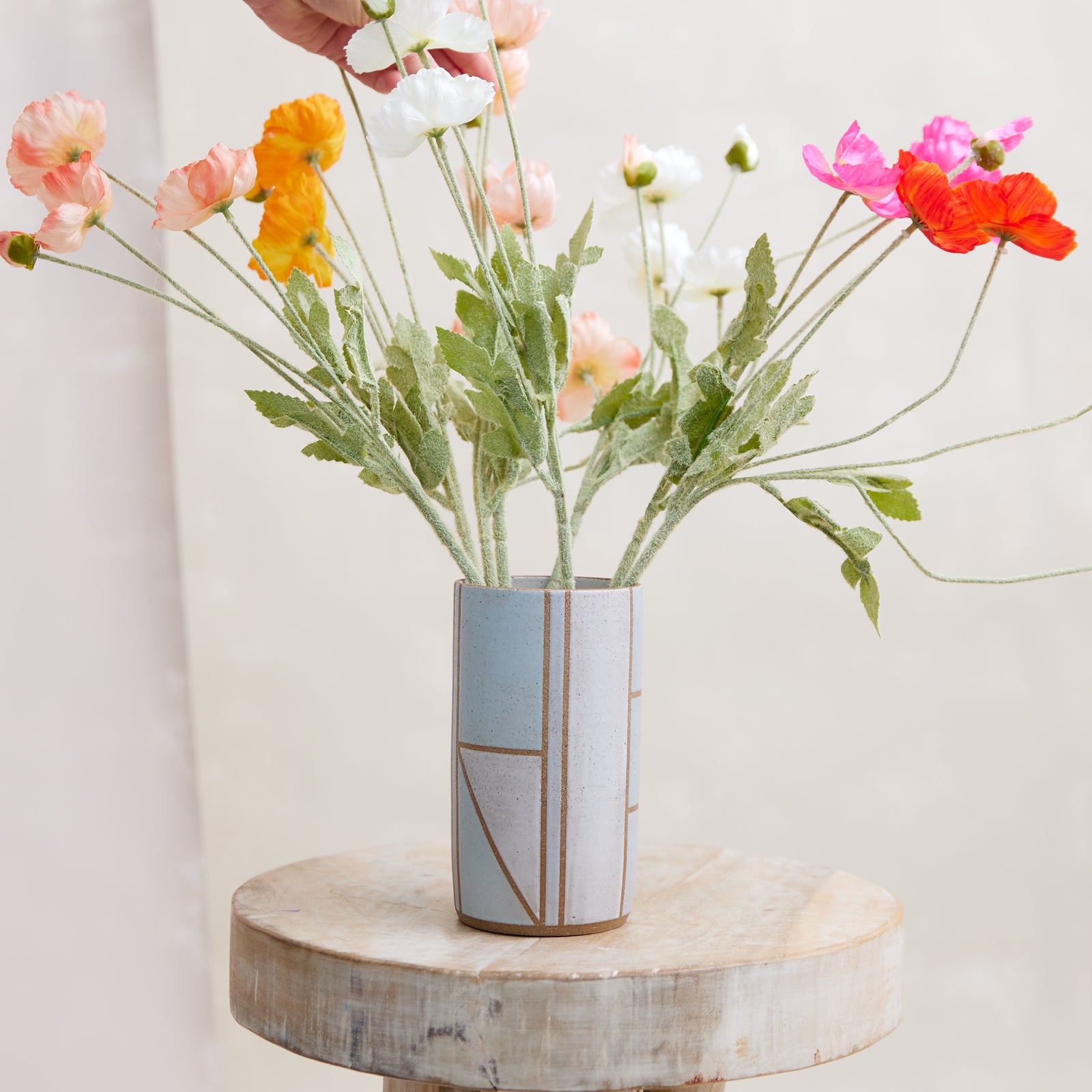 A front view of the Geometric Cylindrical Handmade Ceramic Vase in a blue, white and grey glaze. The handmade vase sits on a wooden stool in a coastal-styled setting. The ceramic vase displays fresh flowers that are being adjusted by a hand.