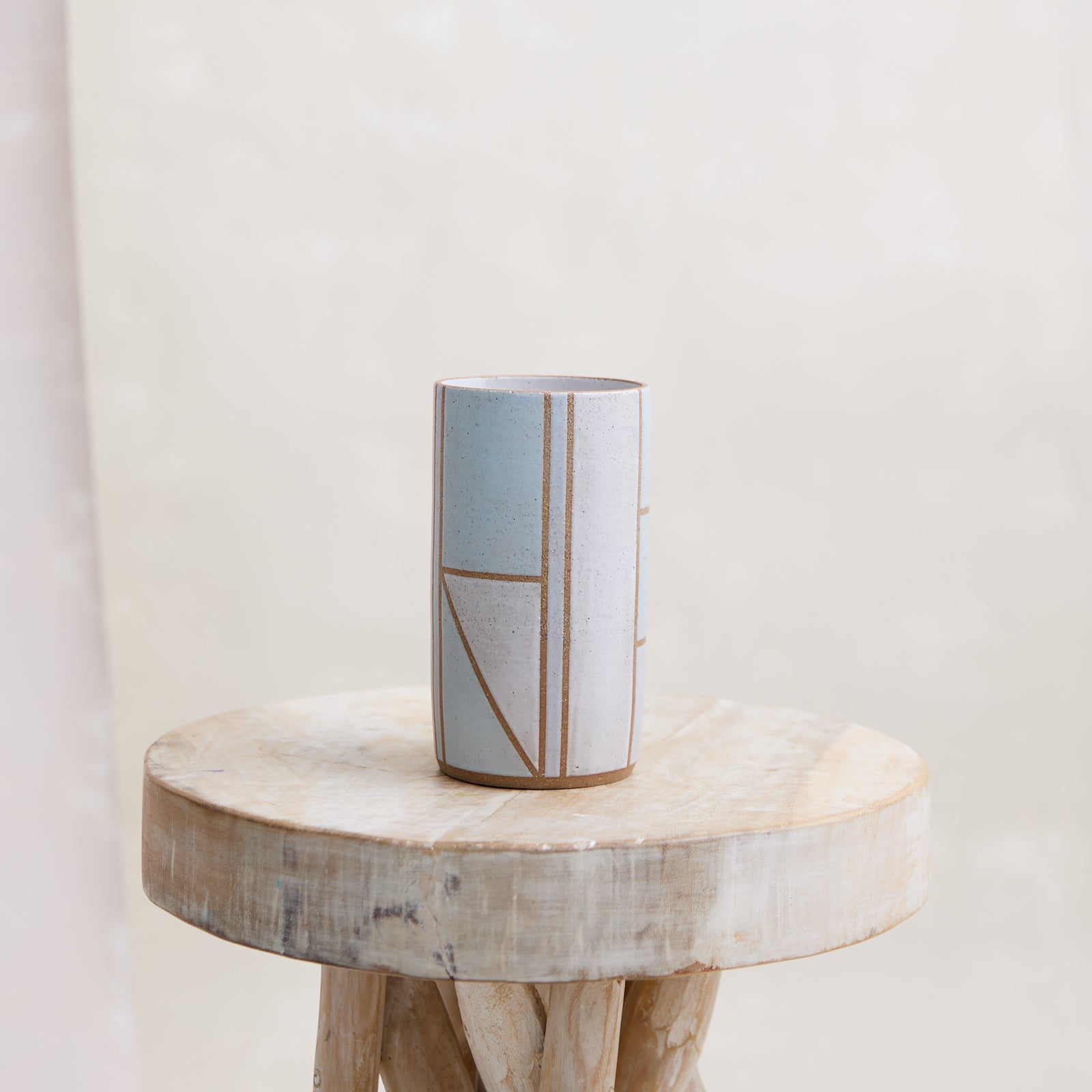 A front view of the Geometric Cylindrical Handmade Ceramic Vase in a blue, white and grey glaze. The handmade vase sits on a wooden stool in a coastal-styled setting. The vase has is cylindrical with a geometric design.