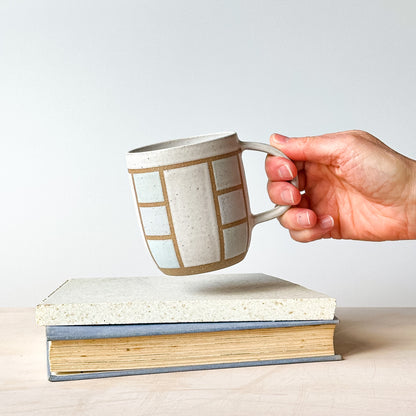 A hand holding a geometric styled mug above a tile.