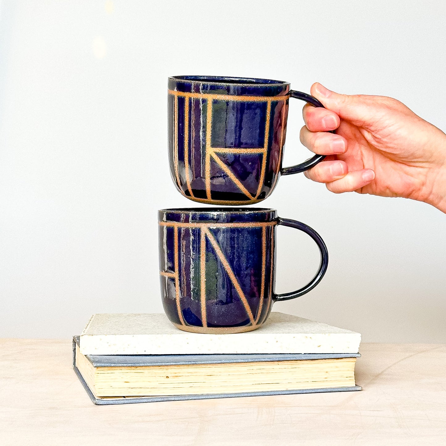 Two ceramic mugs with geometric patterns stacked on books, one held by a hand against a white background.
