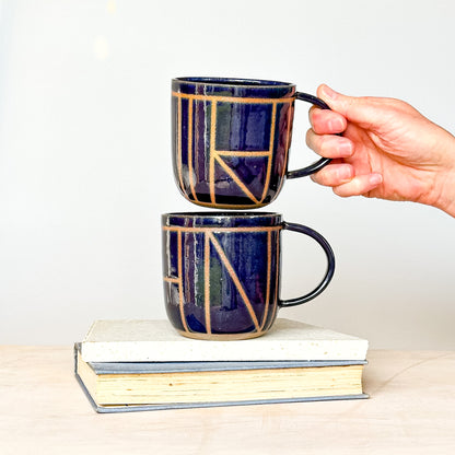 Two ceramic mugs with geometric patterns stacked on books, one held by a hand against a white background.