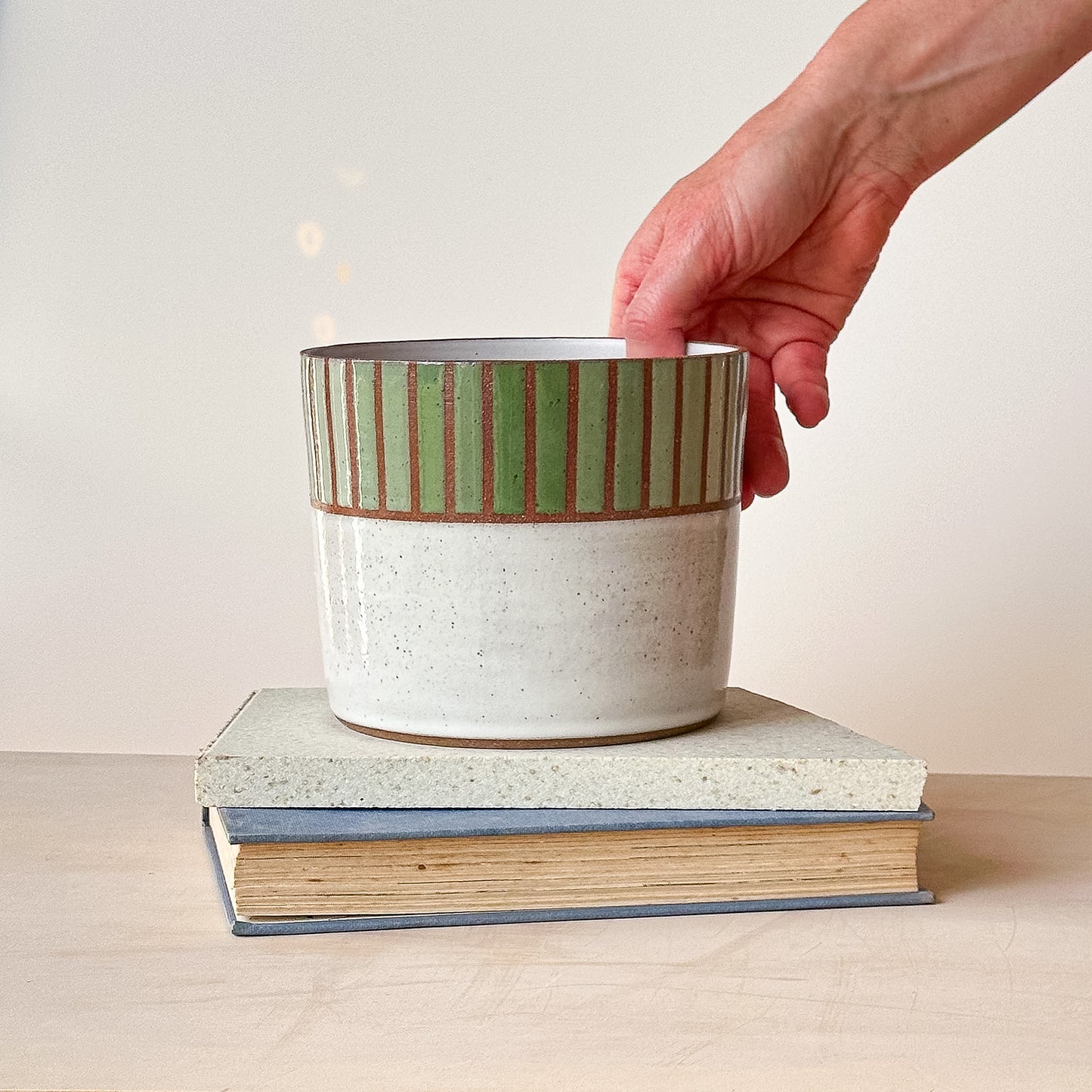 Hand placing a striped ceramic pot on top of two books with a plain background