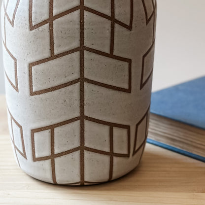 A close up view of a Handmade Geometric Vase. The Vase is glazed white, and the unglazed clay makes the pattern. The handmade vase sits next to a book on a wooden table.