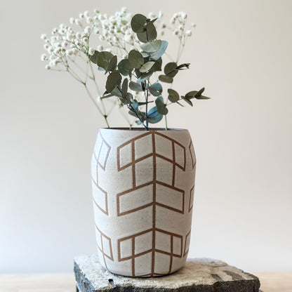 A Geometric Handmade Ceramic vase sits on a tile and on top of a table. The Geometric Ceramic Vase is glazed white. The pattern is made by the unglazed clay. The vase contains an arrangement of gum leaves and baby's breath.