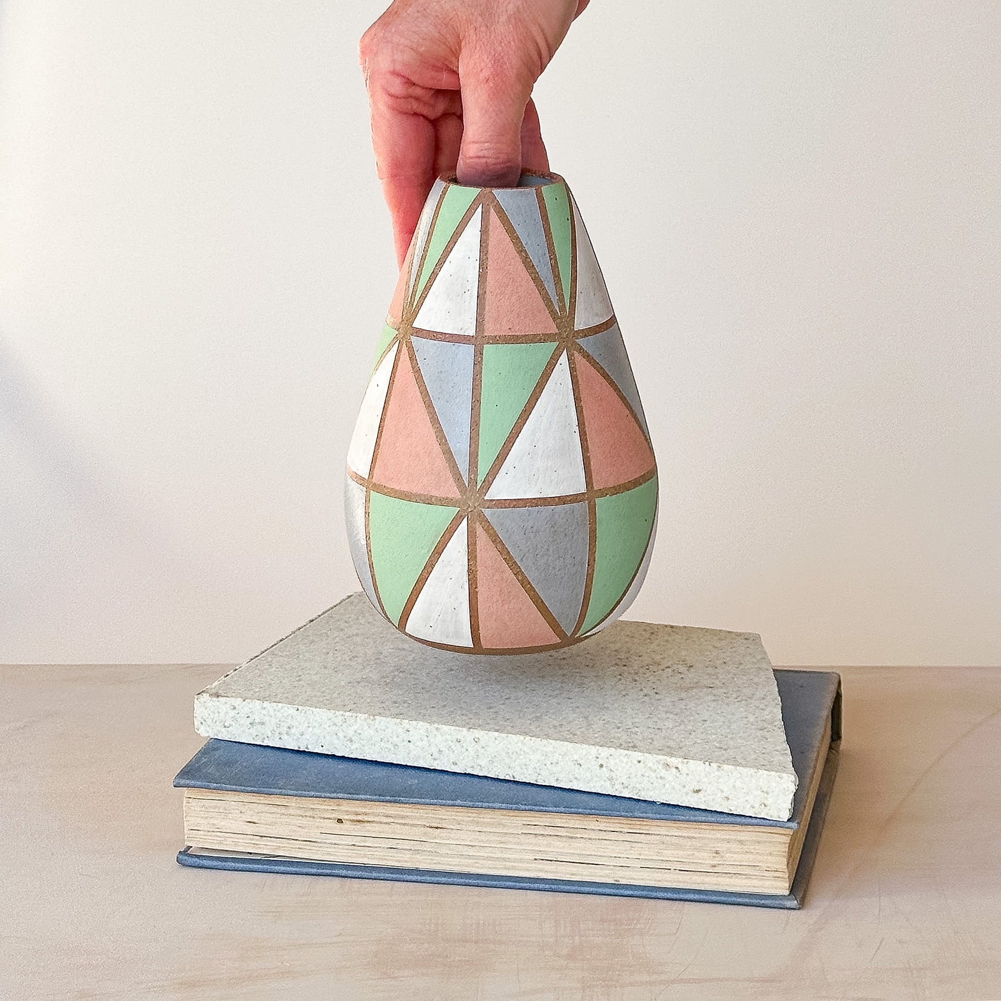 Hand holding a geometric-patterned vase on a stack of books with a plain background