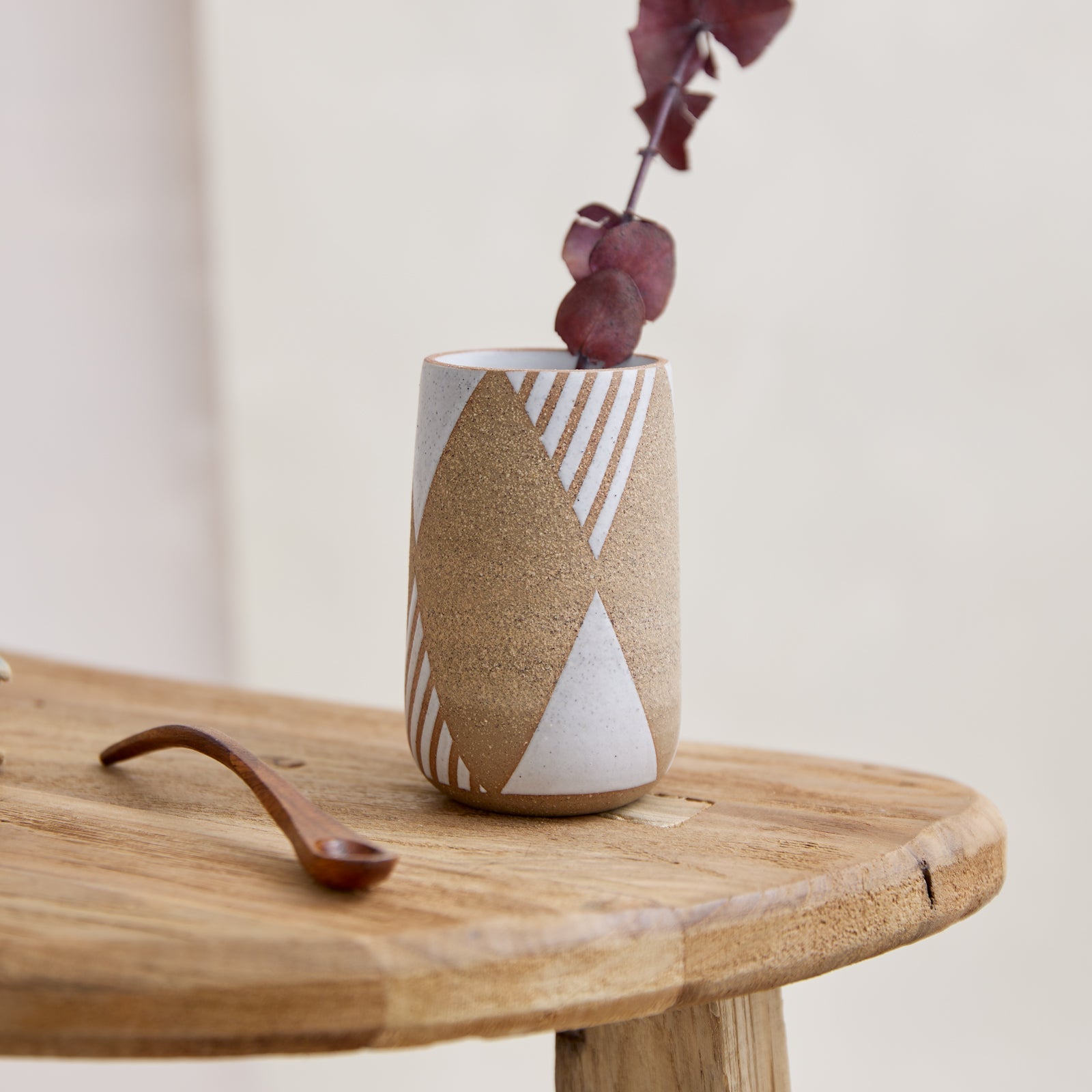  A front view of the Geometric Handmade Ceramic Mini Vase in natural and white glaze. The handmade vase has a geometric design and a tapered opening. The ceramic vase sits on a wooden side table holding flowers in a coastal-styled setting.