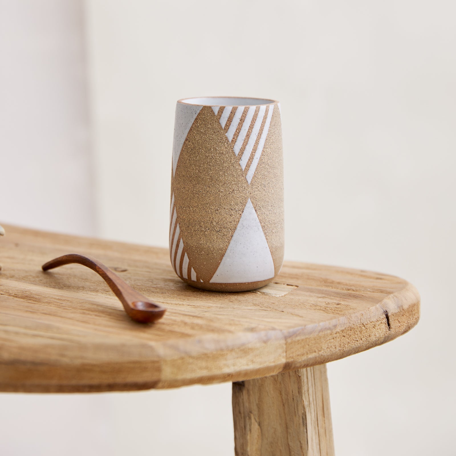  A front view of the Geometric Handmade Ceramic Mini Vase in natural and white glaze. The handmade vase has a geometric design and a tapered opening. The ceramic vase sits on a wooden side table.
