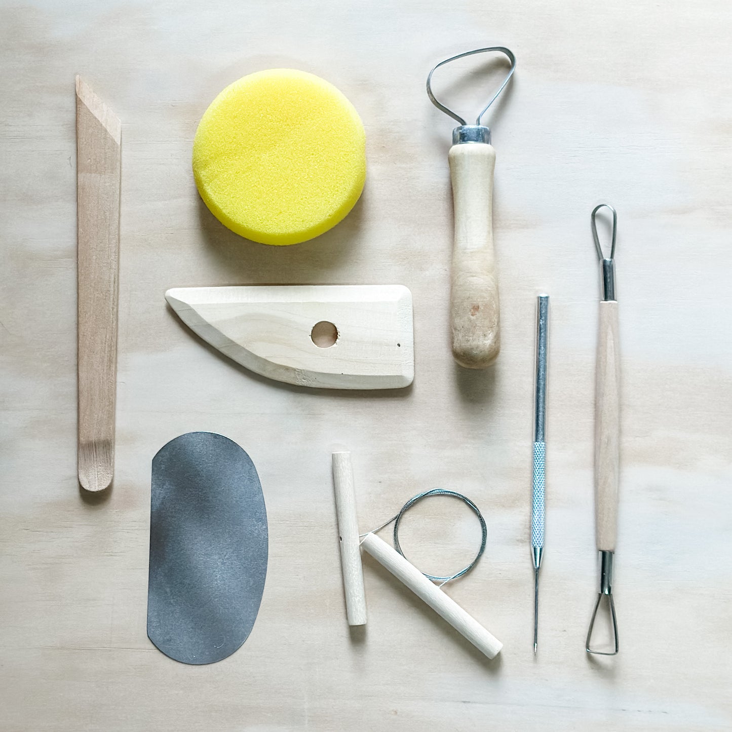 Set of pottery utensils including a yellow sponge, wooden spatula, and metal tools on a light surface.