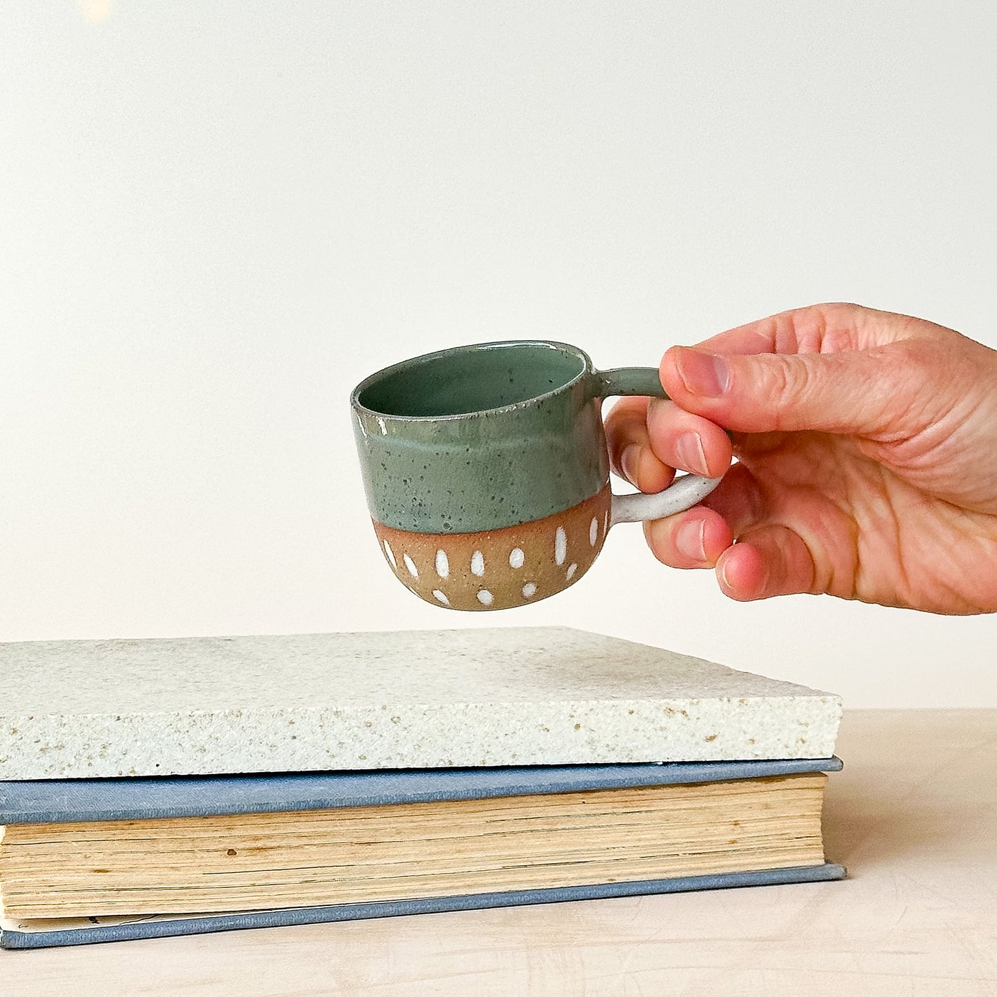 Hand holding a small green ceramic espresso cup over a stack of books on a light background