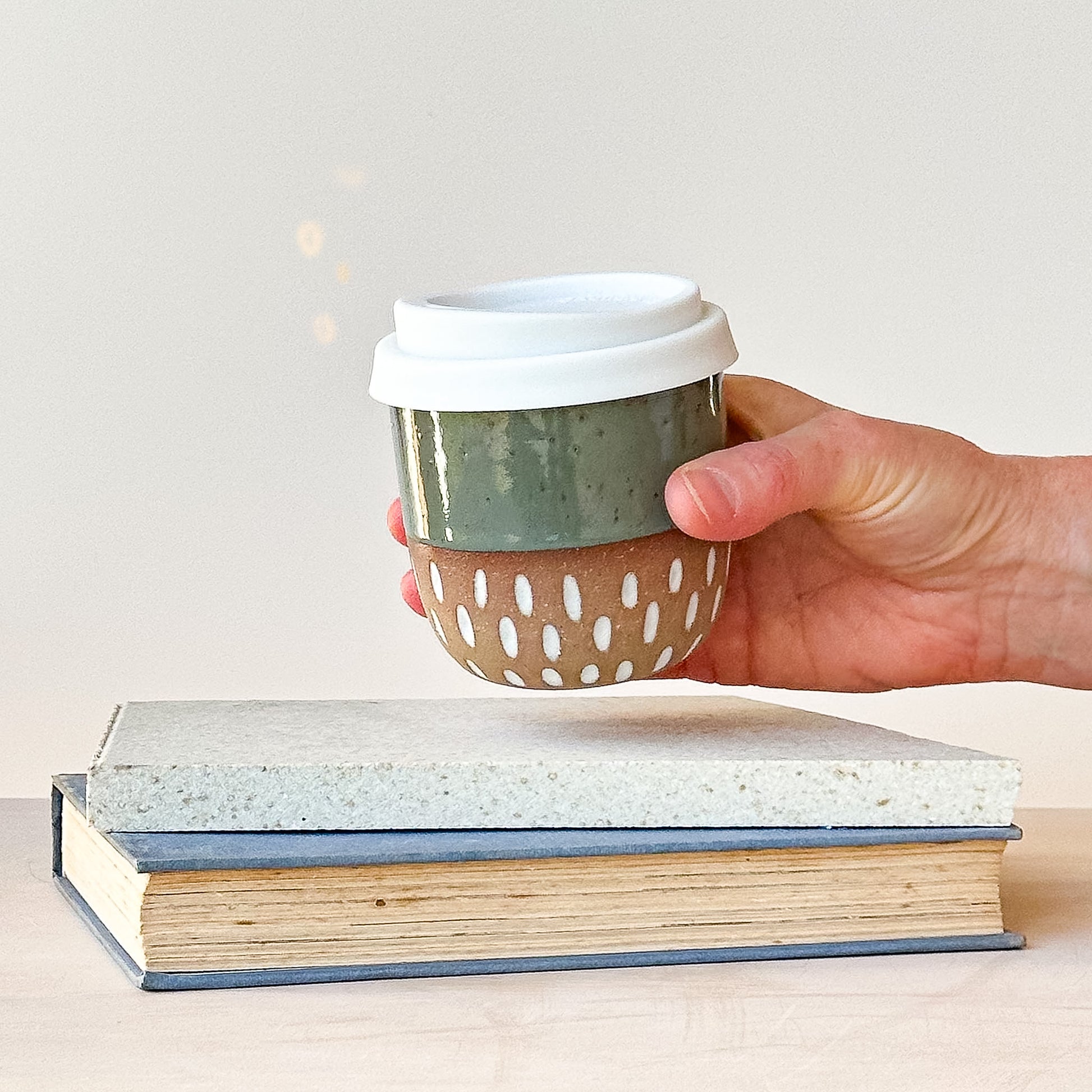Hand holding a ceramic travel mug with a white lid over a stack of books on a light background