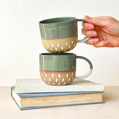 Two ceramic mugs with a textured finish stacked on books, one held by a hand.