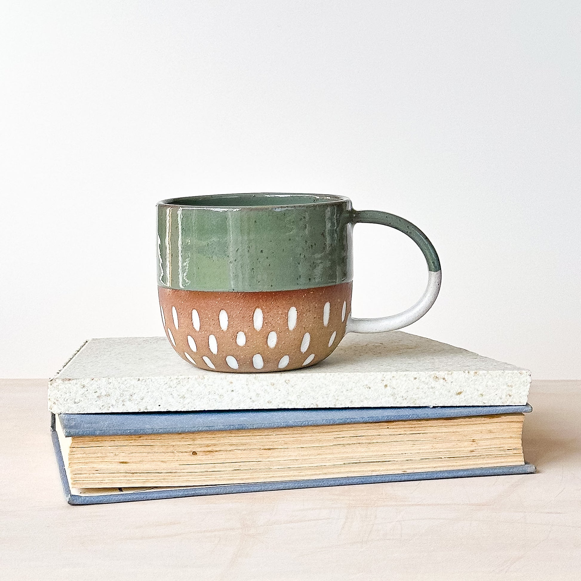 Green and brown ceramic mug with white dots on a stack of books against a light background