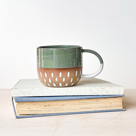 Green and brown ceramic mug with white dots on a stack of books against a light background