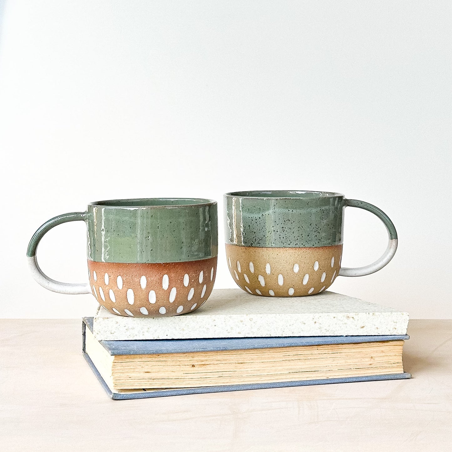 Two ceramic mugs with green tops and brown bottoms on top of books against a white background