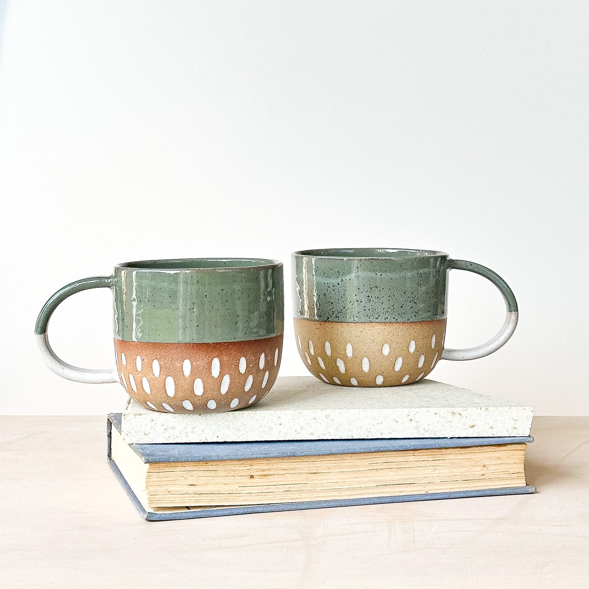 Two ceramic mugs with green tops and brown bottoms on top of books against a white background
