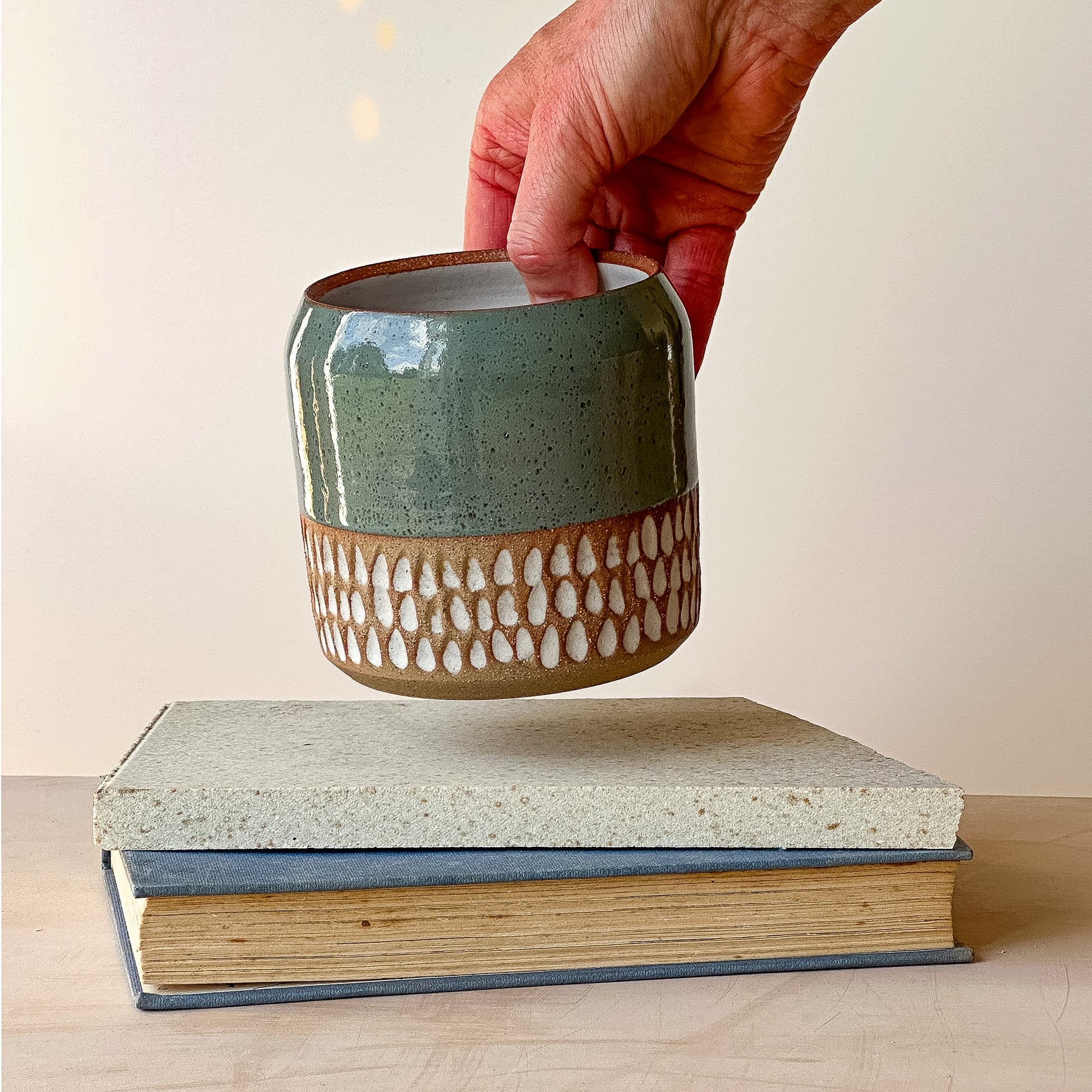 Hand holding a ceramic pot over two stacked books with a plain background