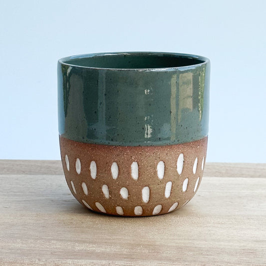 A handmade ceramic cup with a green top and brown bottom with white patterns on a light wooden surface.