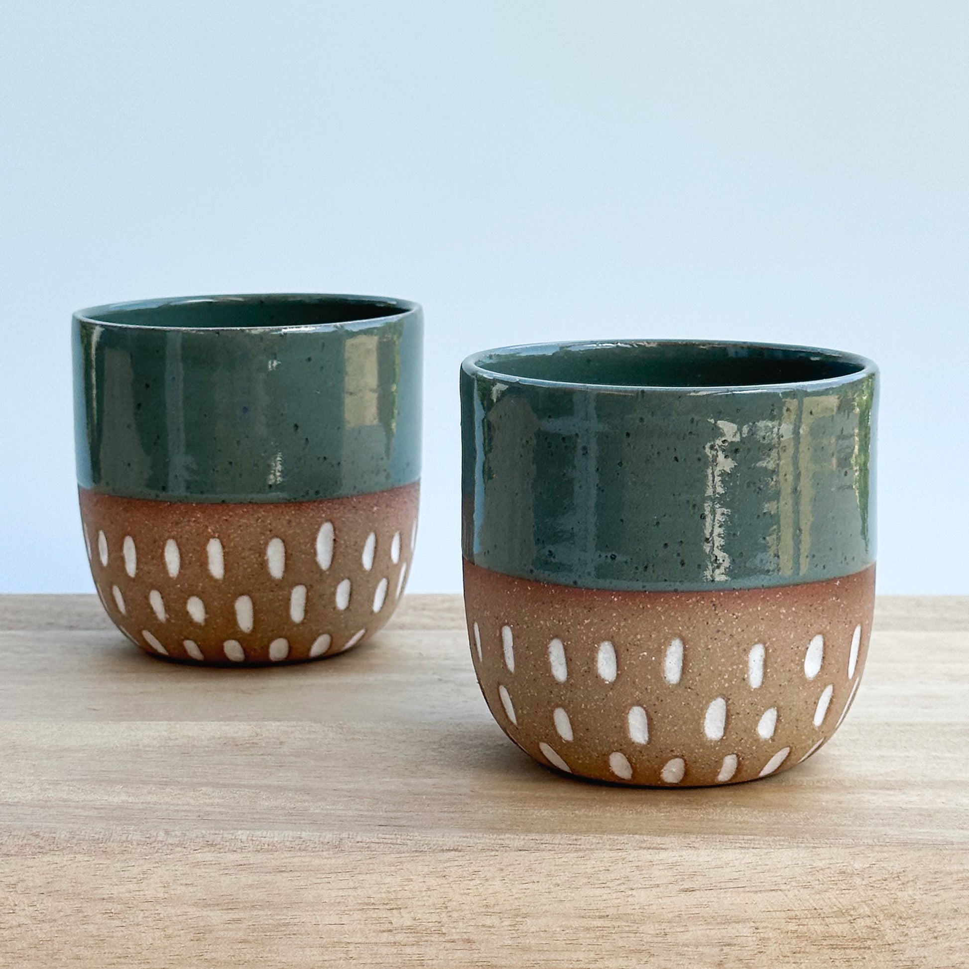 Two ceramic cups with green tops and brown bottoms with white patterns on a wooden surface.