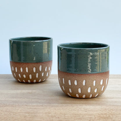 Two ceramic cups with green tops and brown bottoms with white patterns on a wooden surface.