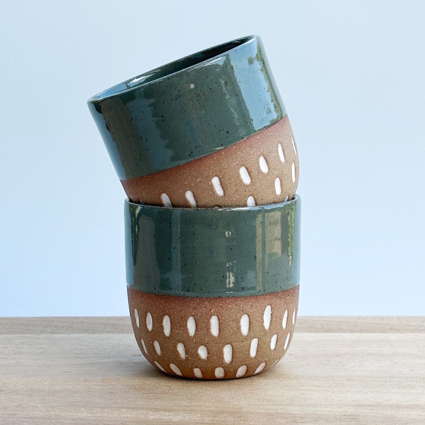 Stack of ceramic tumblers with green and brown design on a light wooden surface and a light background
