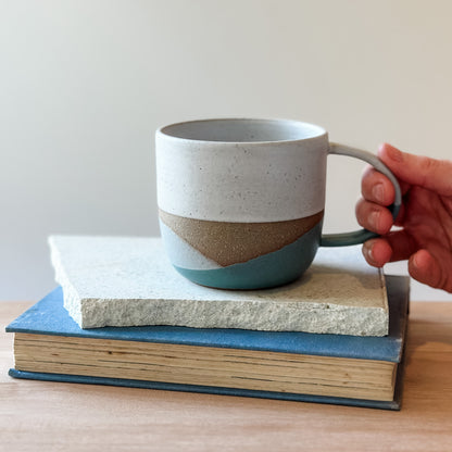 Hand holding a ceramic mug with a coastal design on top of a tile and a book.