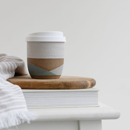 The tidal handmade ceramic keep cup in green and glaze glaze. The handmade ceramic keep cup is sitting on top of a wooden stool in coastal styled setting.