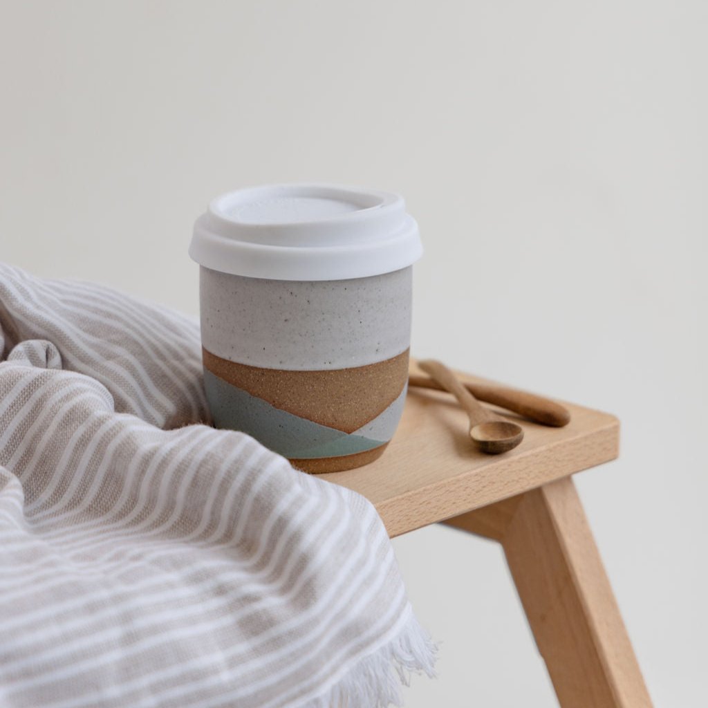 The tidal handmade ceramic keep cup in green and glaze glaze. The handmade ceramic keep cup has a white lid and is sitting on top of a wooden stool in coastal styled setting.