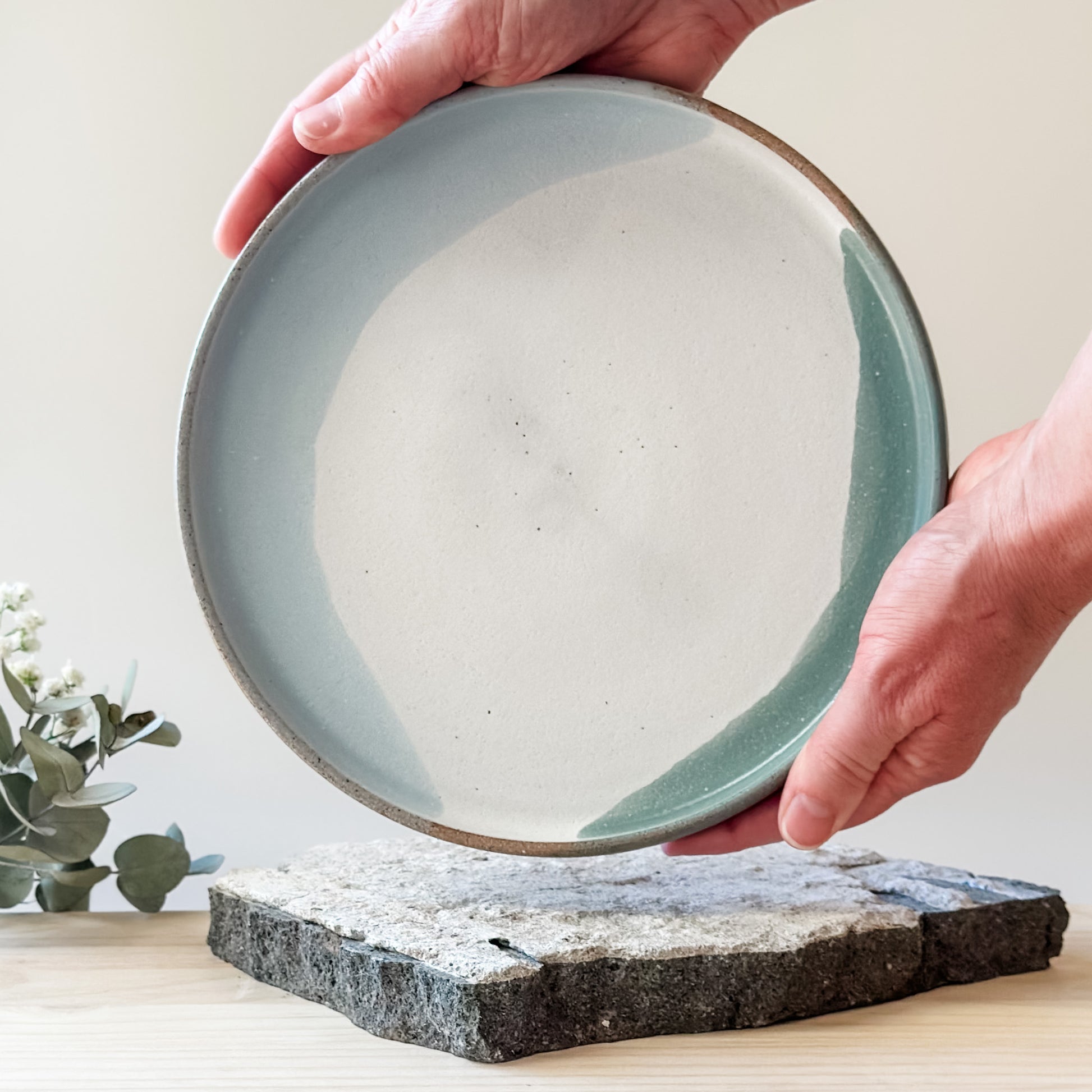 The small Tidal Handmade Ceramic Plate in a white, green and blue-grey glaze. The plate is being held by two hands above a tile and a wooden table.
