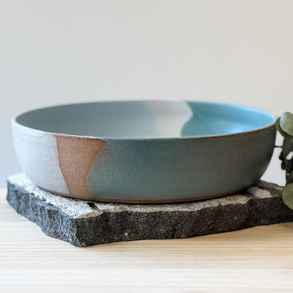 A side view of the Tidal Handmade Ceramic Serving Bowl. The handmade bowl sits on a tile and a wooden table.
