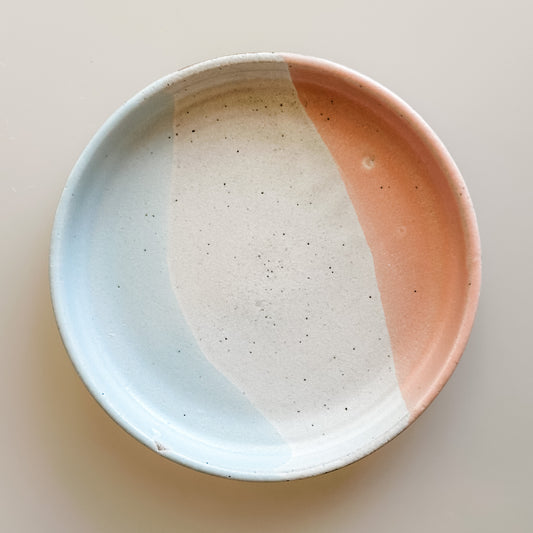 A large handmade ceramic trinket dish sits on a cream background. The dish is glazed white, blue-grey, and peach.