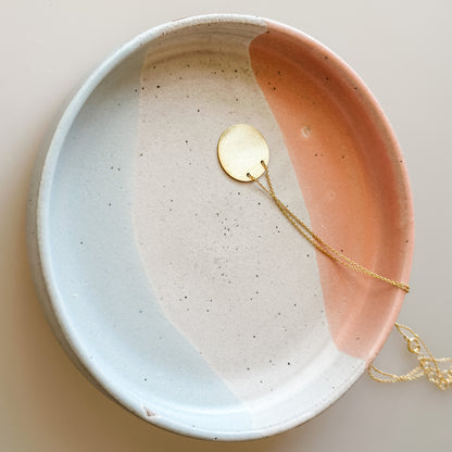 A large handmade ceramic trinket dish sits on a cream background. The dish, which is glazed white, blue-grey, and peach, holds a gold necklace.