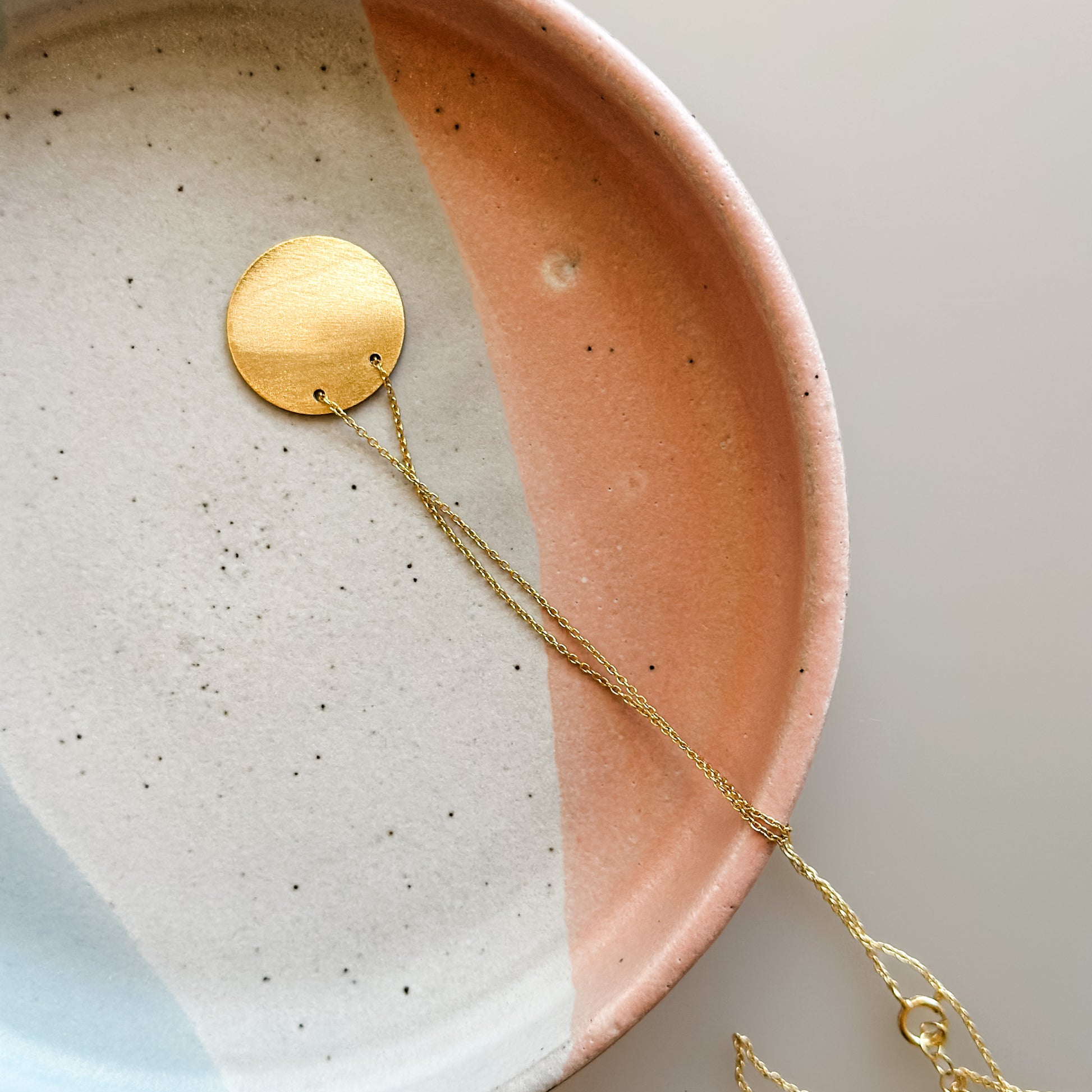 A large handmade ceramic trinket dish sits on a cream background. The dish, which is glazed white, blue-grey, and peach, holds a gold necklace.